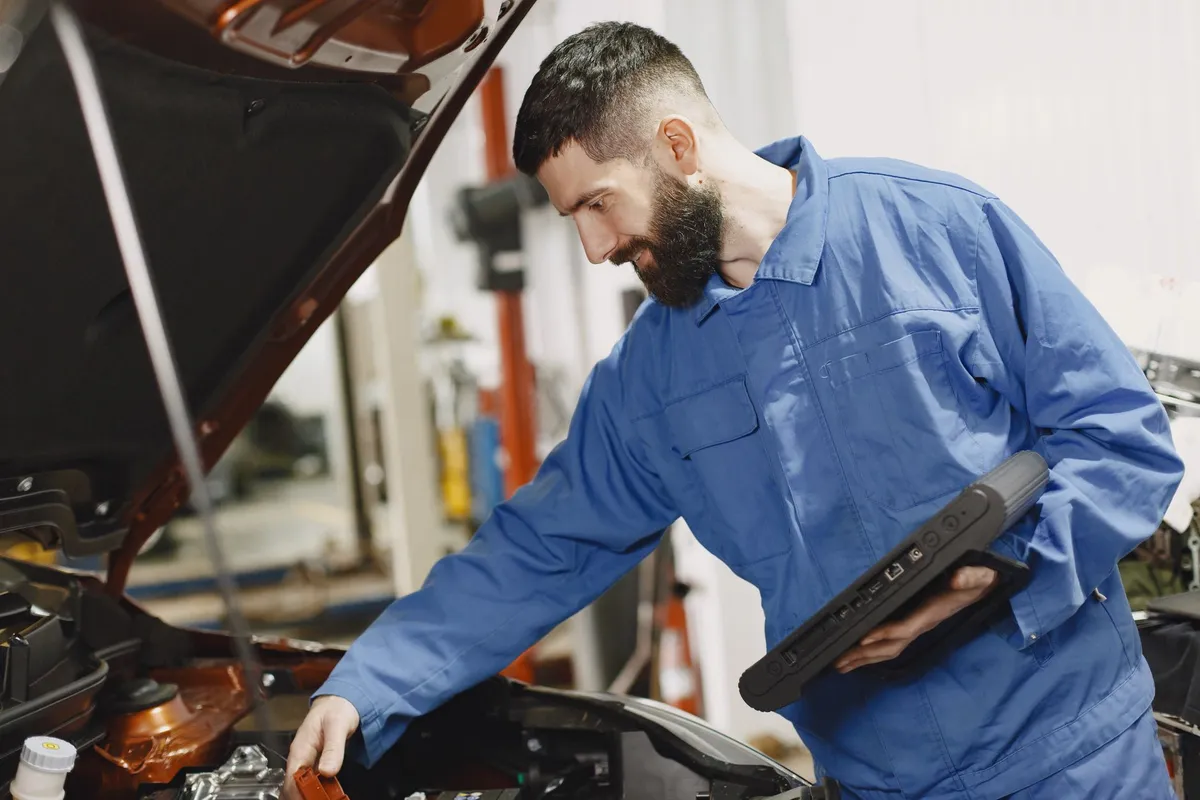 A mechanic examining a car engine bay while holding a diagnostic tablet