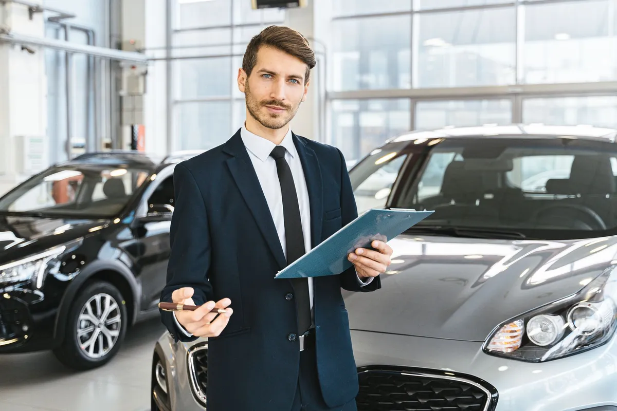 A dealership salesperson holding a clipboard standing between two vehicles in a showroom