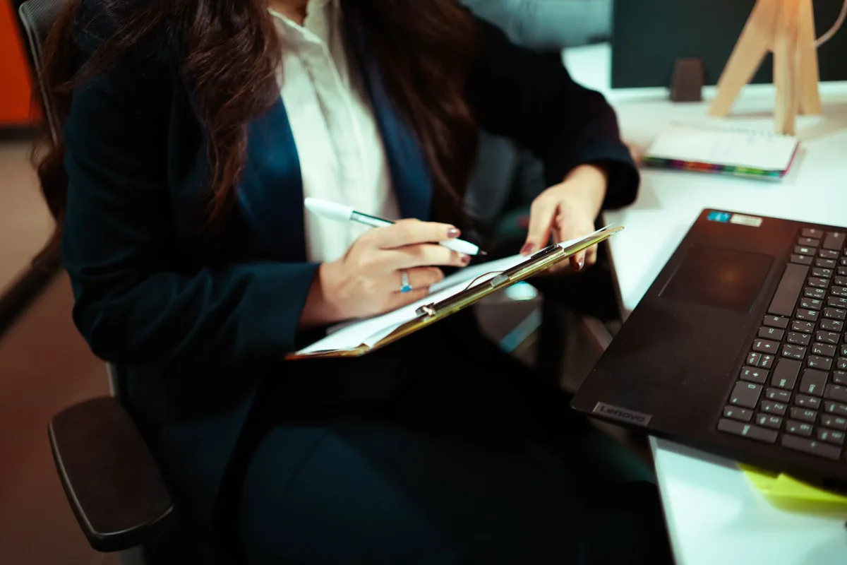 A professional writing on a clipboard at a desk with a laptop
